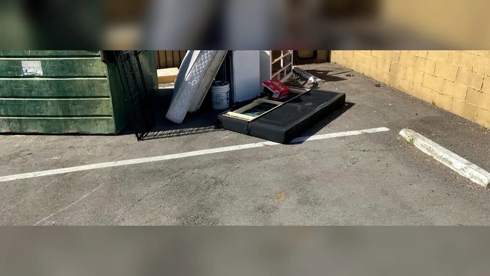 Palmdale parking area before cleanup with a mattress, appliance, treadmill base, and boxes beside a dumpster.